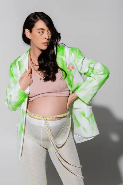 brunette pregnant woman in green and white blazer, crop top, beads belt and leggings holding hand on waist and looking away on grey background, maternity style concept, expectation