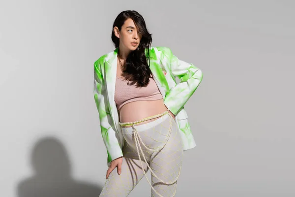 ブルーネット妊娠中の女性で緑と白のジャケット タイツ クロップトップ ビーズベルト立っています手でヒップでグレーの背景 産前スタイルのコンセプト — ストック写真