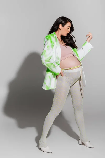 full length of future mother in crop top, stylish blazer, leggings and beads belt standing and holding hand on waist on grey background, expectation, maternity fashion concept