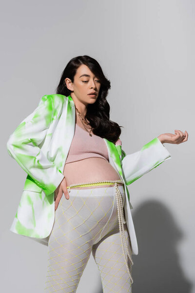 fashionable mom-to-be in green and white blazer, crop top, beads belt and leggings standing with hand on hip on grey background, expectation, pregnancy style concept