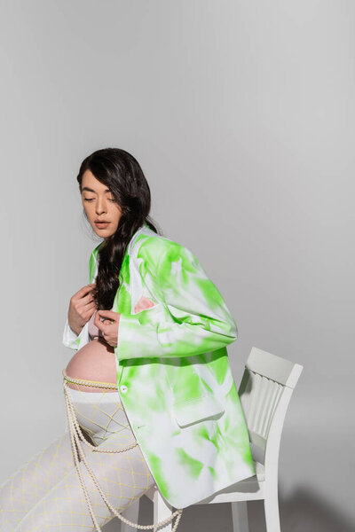 pregnant woman with wavy brunette hair, wearing trendy jacket, crop top, beads belt and leggings, sitting on chair on grey background, maternity fashion concept, expectation