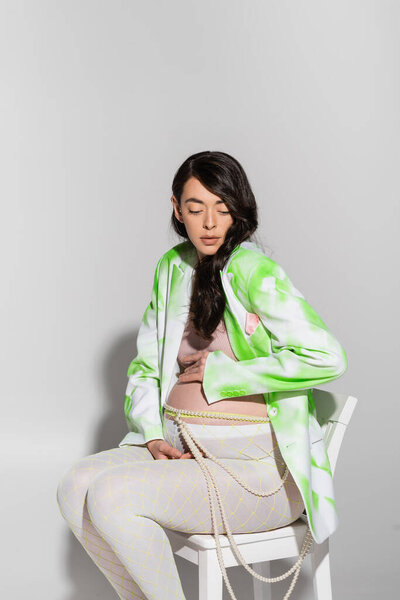 brunette pregnant woman in stylish blazer, crop top, beads belt and leggings sitting on chair and touching tummy on grey background, maternity fashion concept, expectation