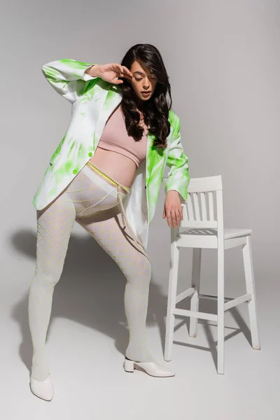 緑と白のブレザーで魅力的な妊婦の完全な長さ 作物の上 グレーの背景に椅子の近くにポーズビーズベルトとレギンス ファッショナブルな妊娠の概念 — ストック写真