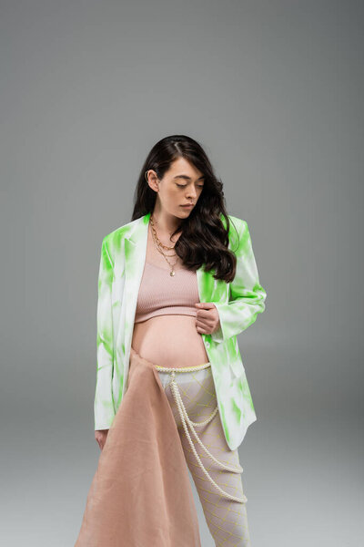 brunette pregnant woman in green and white blazer, crop top, beads belt and leggings with beige chiffon cloth on grey background, maternity fashion concept