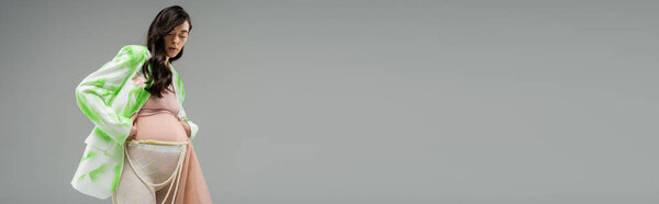 low angle view of future mother in trendy green and white blazer, crop top, leggings with beads belt and chiffon cloth isolated on grey background, pregnancy fashion concept, banner