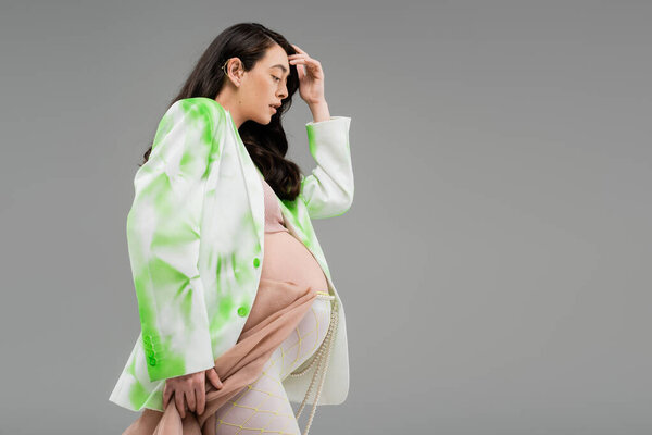side view of pretty pregnant woman in green and white jacket and leggings with beads belt and beige chiffon cloth isolated on grey background, maternity fashion concept, expectation