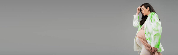 side view of pregnant woman with wavy brunette hair posing in green and white blazer, crop top and leggings with chiffon cloth and beads belt isolated on grey background, banner