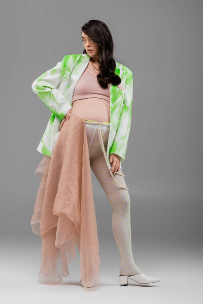 full length of brunette pregnant woman in green and white blazer, crop top and leggings with beige chiffon cloth and beads belt standing with hand on hip on grey background, maternity fashion concept