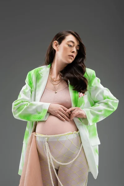 appealing future mother in green and white jacket, crop top, beads belt and tights with beige chiffon cloth isolated on grey background, expectation, maternity fashion concept