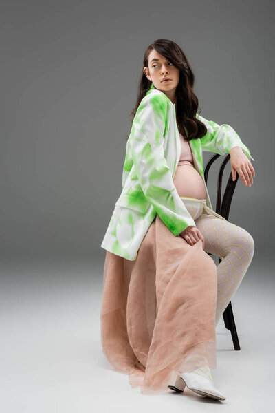 full length of brunette mother-to-be in stylish jacket and leggings with beads belt and beige chiffon cloth sitting on chair and looking away on grey background, fashionable pregnancy concept