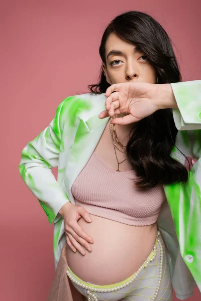 brunette pregnant woman in crop top, fashionable blazer and beads belt holding hand near face, touching tummy and looking at camera isolated on pink background, maternity style concept