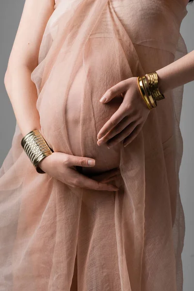 怀孕妇女穿着柔和的雪纺窗帘和金色的手镯 抱着灰色背景的肚子 生育时尚观念 未来的腹部母亲 — 图库照片