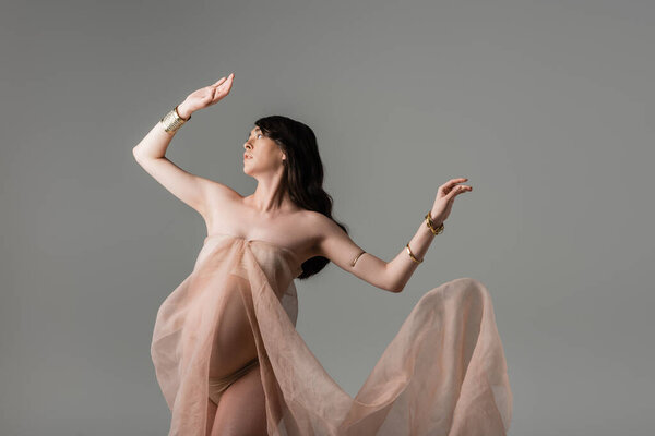 charming mother-to-be in flowing chiffon draping and golden accessories posing and looking away isolated on grey background, maternity fashion concept, pregnant woman with belly 