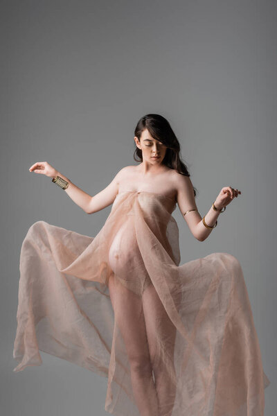 elegant expecting mother with wavy brunette hair posing in delicate chiffon draping and golden bracelets isolated on grey background, maternity fashion concept, pregnant woman with belly 