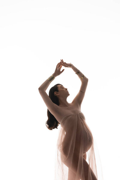 future mother in golden bracelets and transparent chiffon cloth posing with closed eyes and raised hands isolated on white background, maternity style concept, expectation, woman with belly
