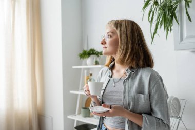 Genç bir kadın, kısa saçlı, kâhküllü, gözlüklü ve dövmeli, elinde kahve fincanı ve çay tabağı tutarken, modern mutfakta bulaşık rafının ve bitkinin yanında gri günlük giysiler içinde duruyor. 
