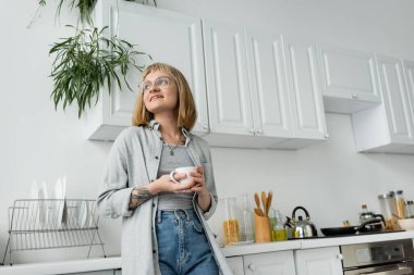 joyful young woman with short hair and bangs, eyeglasses and tattoo holding cup of morning coffee while looking away and standing in casual clothes next to dishes, kettle, kitchen appliances  clipart