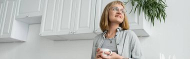 positive young woman with short hair and bangs, eyeglasses and tattoo holding cup of morning coffee while looking away and standing in casual clothes next to white kitchen cabinet and plant, banner  clipart