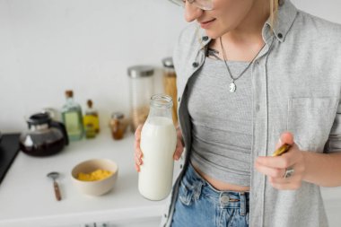 cropped view of tattooed woman holding bottle with fresh milk near blurred bowl with cornflakes and spoon while making breakfast and standing in casual clothes next to kitchen appliances at home  clipart