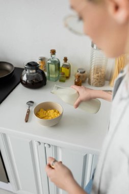 blurred and tattooed young woman in eyeglasses holding bottle while pouring fresh milk into bowl with cornflakes on kitchen worktop while making breakfast and standing in modern kitchen  clipart