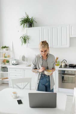Kahkahalı, gözlüklü, kısa saçlı, kahvaltıda mısır gevreğiyle kase tutan, akıllı telefonun yanında dizüstü bilgisayara bakan ve modern mutfakta masanın üstünde kahve içen dövmeli genç bir kadın. 