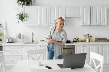 tattooed young freelancer with bangs and eyeglasses smiling, holding bowl with cornflakes while having breakfast and looking at laptop near smartphone and cup of coffee on desk in modern kitchen  clipart