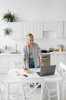 tattooed freelancer with bangs and eyeglasses holding bowl with cornflakes while having breakfast and looking at laptop near smartphone and cup of coffee on saucer and desk in modern kitchen  clipart