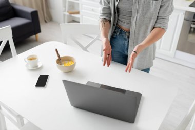 Akıllı telefonun yanında elinde dövmeli genç bir kadın, boş ekranlı bir laptop, mısır gevrekli bir kase, modern mutfağın masasındaki beyaz çay tabağında bir fincan kahve ve bir fincan kahve.