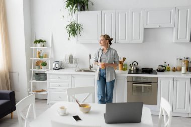 tattooed woman with bangs and eyeglasses holding glass of orange juice and standing near kitchen worktop next to desk with devices, bowl with cornflakes and cup of coffee on saucer at home, freelance  clipart
