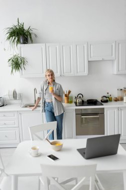 tattooed woman in eyeglasses holding glass of orange juice and standing near desk with devices, bowl with cornflakes and cup of coffee with saucer on desk in modern kitchen, looking at laptop  clipart