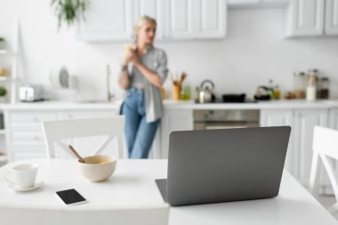 Bilgisayar, boş ekranlı akıllı telefon, mısır gevrekli kase, beyaz masadaki fincan tabağı, modern mutfakta duran bulanık serbest çalışan, serbest çalışan, dijital göçebe. 