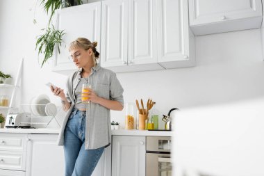 tattooed woman with bangs and eyeglasses holding glass of orange juice and using smartphone while standing near clean dishes and green plants in blurred white kitchen in modern apartment  clipart