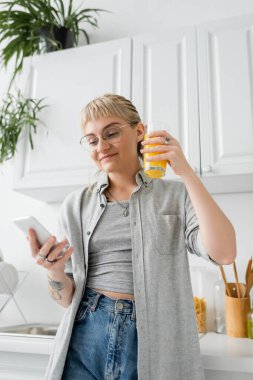tattooed and happy woman with bangs and eyeglasses holding glass of orange juice and using smartphone while standing near clean dishes and blurred green plants in modern apartment  clipart