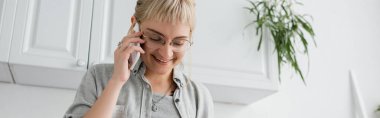Kâküllü ve gözlüklü mutlu genç bir kadın akıllı telefonuyla gülümseyip konuşurken, gözleri kapalı bir şekilde modern dairede bulanık yeşil bitkiler ve beyaz mutfak dolaplarının yanında duruyor.