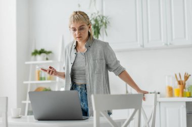 Kısa saçlı, dövmeli ve kâküllü genç bir kadın akıllı telefon kullanırken bir fincan kahve bardağının yanında duruyor ve modern apartmandaki sandalyelerin etrafındaki bulanık laptopa bakıyor. 