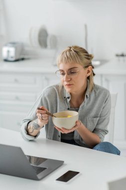 young woman with bangs and tattoo on hand eating cornflakes for breakfast while looking at laptop near smartphone with blank screen on table in modern kitchen, freelancer, work from home  clipart