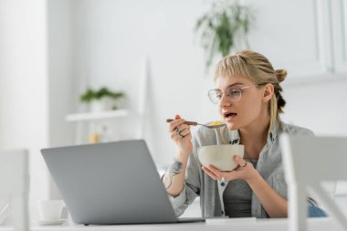 Kahküllü ve dövmeli genç bir kadın kahvaltıda mısır gevreği yerken akıllı telefonun yanında dizüstü bilgisayar ve modern mutfaktaki masada bir fincan kahve kullanıyor. 