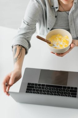 Elinde dövmesi olan genç bir kadın kahvaltıda mısır gevreğiyle kaseyi tutan gri gömlekli ve modern dairede beyaz masada dizüstü bilgisayar kullanan serbest çalışan, üst görüşlü bir kadın. 