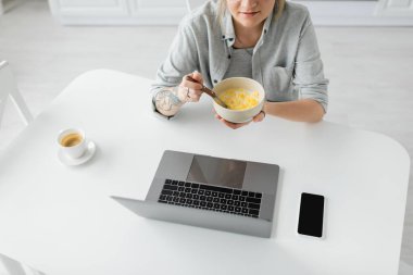 Kahvaltıda elinde dövmesi olan genç bir kadının mısır gevreği yerken akıllı telefonun yanında boş ekranlı dizüstü bilgisayar ve modern mutfaktaki kahve fincanı kullanırken çekilmiş görüntüsü. 