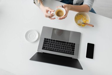 Kahvaltı sırasında elinde dövmesi olan bir kadının elinde mısır gevreğiyle kasenin yanında kahve fincanı tutarken modern mutfakta boş ekranlı akıllı telefon, serbest çalışan, kesilmiş... 