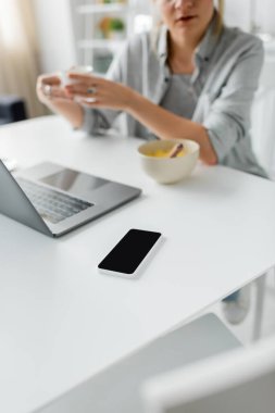 Kahvaltıda elinde mısır gevreğiyle kasenin yanında bulanık kahve tutan bir kadının modern mutfakta boş ekranı olan akıllı telefonun yanında dizüstü bilgisayar kullanırken görüntüsü. 