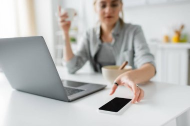Kahvaltıda elinde mısır gevreğiyle kasenin yanında kahve bardağını tutan bulanık genç bir kadın dizüstü bilgisayar kullanırken modern mutfakta boş ekranla akıllı telefona uzanıyor. 
