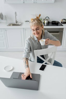 Elinde dövmesi olan, elinde kahve fincanı olan, akıllı telefon ve çay tabağının yanındaki dizüstü bilgisayara bakan, modern mutfakta sandalyelerin etrafında uçan dairelere bakan mutlu genç bir kadın.