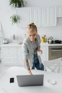 Yüksek açılı, elinde dövmesi olan, elinde kahve fincanı olan ve bilgisayarını akıllı telefon ve tabağın yanında kullanan modern mutfakta, serbest çalışan, uzak yaşam tarzı olan genç bir kadın. 