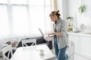 Gözlüklü, kısa saçlı ve kâküllü genç bir kadın bir fincan kahve ve akıllı telefonun yanında dizüstü bilgisayar tutuyor beyaz masalarda beyaz ve modern mutfaktaki sandalyelerin etrafında kanepe, serbest çalışan, uzak yaşam tarzı olan