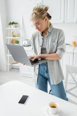 Gözlüklü, kısa saçlı ve kâküllü genç bir kadın bir fincan kahvenin yanında dizüstü bilgisayar ve beyaz ekranlı akıllı bir telefon beyaz masadaki beyaz mutfak, uzaktan yaşam tarzı, serbest çalışan bir kadın.