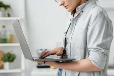 Beyaz ve modern mutfakta dizüstü bilgisayar tutan, kullanan, gri tişörtlü genç bir kadının görüntüsü bulanık arka plan, uzak yaşam tarzı, serbest çalışan, evden çalışan, serbest çalışan...