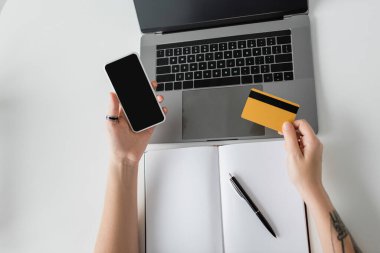Kredi kartı ve akıllı telefon tutan dövmeli kadının üst görüntüsü dizüstü bilgisayarın yanında boş ekran ve beyaz masa üzerinde kalemli defter, modern çalışma alanı, evden çalışma, dijital ödeme  