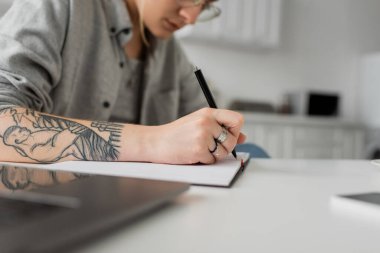 Eline dövme yaptırmış genç bir kadının not defterine yazdığı, not aldığı, kalemini beyaz masada tutarken ilham aldığı, ön planda bulanıklık olan, evden iş yaptığı görüntüler. 