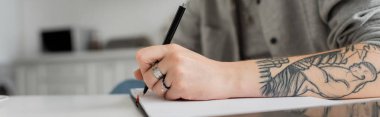 cropped shot of young woman with tattoo on hand writing in notebook, taking notes, having inspiration while holding pen near laptop on white table, blurred foreground, work from home, banner  clipart
